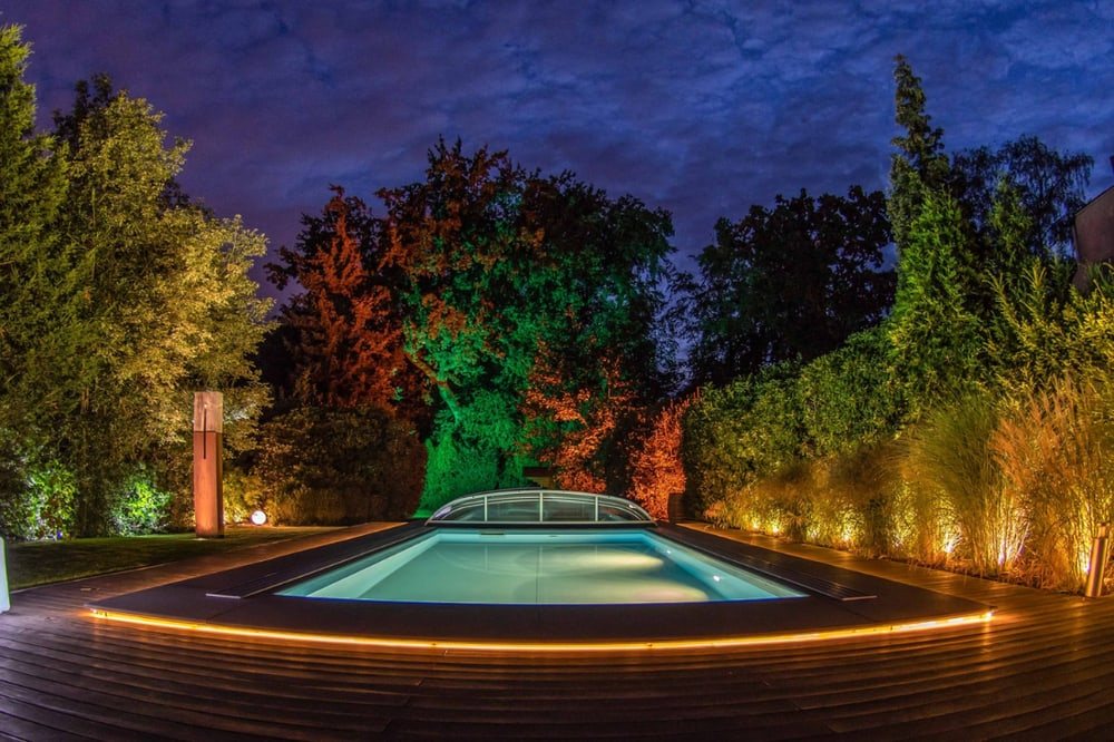 Beleuchteter Pool bei Nacht, umgeben von Holzdielen und Bäumen